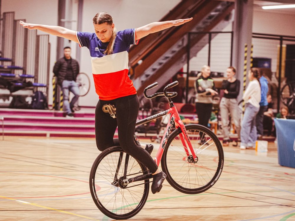 Une cycliste en maillot de l'équipe de France, debout en équilibre sur son vélo de cyclisme artistique, bras écartés, dans un gymnase.